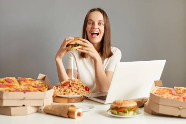 Aşırı mutlu kadın dizüstü bilgisayarının portresi bilgisayar ve fast food ile masada oturuyor, keyifli bir son diyet yapıyor, abur cubur yiyor, gri arka planda izole edilmiş.