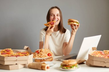 Doymuş bir kadının portresi dizüstü bilgisayarı ve farklı sağlıksız yiyeceklerle dolu gri arka planda izole edilmiş pizza yiyor, abur cuburların tadını çıkarıyor..