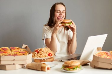 Sinema dizüstü bilgisayarı seyreden aç bir kadının portresi, dinlenirken hamburger yiyen, gri arka planda izole edilmiş aldatılmış bir yemek yiyen, dilinin dışarı çıktığını gösteren..