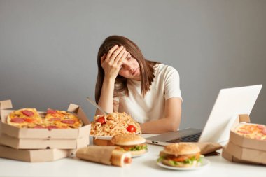 Dizüstü bilgisayarda çalışan, geniş bir fast food çeşitliliğiyle çevrili, alnında avuç içi kadar olan, gri arka planda izole edilmiş, aşırı çalışkan kadının portresi..