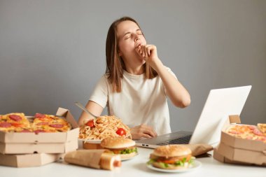 Uzun saatler boyunca dizüstü bilgisayarda çalışan ya da film izleyen uykulu kadının görüntüsü gri arka planda izole edilmiş pizza, makarna, sosisli sandviç ve hamburgerlerle çevrili..