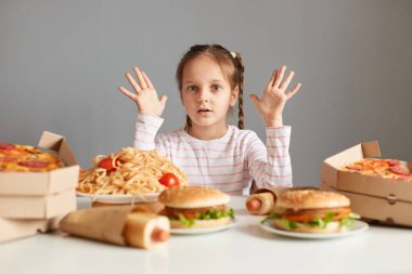 Şaşırmış küçük bir kızın yemek yerken, fast food dağıtırken, büyük yemek takımlarının arasında masada otururken, pizza, hamburger, makarna gri arka planda izole edilmiş, büyük gözlerle kameraya bakarken..