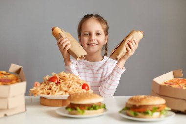 Gri arka plan üzerinde izole bir masada oturan, elinde sosisli sandviç tutan, sahte yemek yiyen, mutlu yüzlü kameraya bakan mutlu, küçük, şirin bir kız resmi..