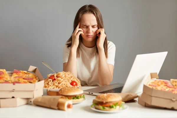 在垃圾食品中坐在笔记本电脑前的生病的不健康妇女的室内照片 她头疼得可怕 疲惫不堪 被灰色背景隔离 — 图库照片