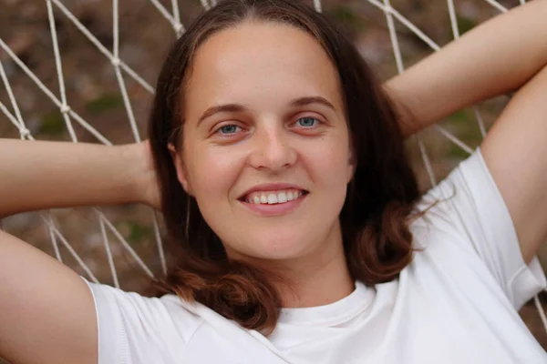 Closeup portrait of young woman with brown hair wearing casual white t-shirt lying in hammock outdoors, raised her arms behind head, looking at camera, relaxing in open air.