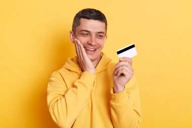 Çevrimiçi bankacılık. Kredi kartı ödemesi. Sıradan bir kapüşonlu giyen, mutlu ve çekici bir adam. Banka kartı tutuyor. Sarı arka planda izole edilmiş pozlar için ödeme yapıyor..