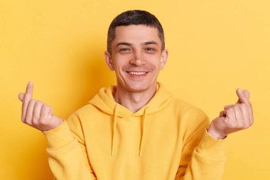Küçük jestlere bayılırım. Gülümseyen mutlu romantik adam, günlük kapüşonlu, Kore aşk işareti gösteren, sarı arka planda izole edilmiş pozlarla kameraya bakan..