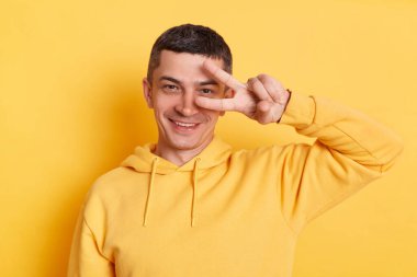 Neşeli, yakışıklı, günlük kapüşonlu bir adam zafer işareti gösteriyor. Kameraya gülümsüyor, sarı arka planda izole edilmiş pozitifliği ifade ediyor..