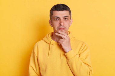Düşünceli, düşünceli, kapüşonlu, sıradan bir adam. Çenesini tutuyor, kamera planına bakıyor. Sarı arka planda izole edilmiş poz vermeyi düşünüyor..