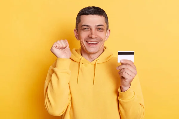 stock image Online shopping. Banking paying. Delighted cheerful man wearing casual hoodie earning big sum of money clenched fist celebrating succes posing isolated over yellow background.