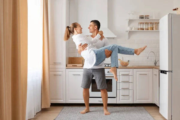 一对年轻夫妇在现代厨房里摆出一副笑脸 牵着他的妻子 享受着穿着白衬衫和牛仔裤的温馨时光 — 图库照片