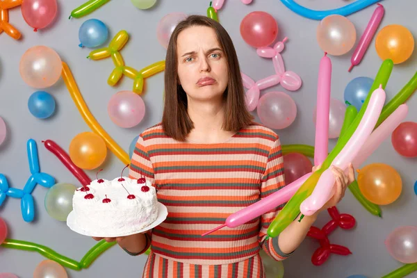 Upset disappointed woman with brown hair wearing striped dress holding cake being upset has bad mood looking with pout lips standing against gray wall decorated with colorful balloons.