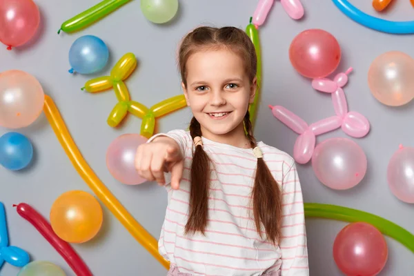I need you. Smiling cute little girl  with pigtails pointing to camera inviting you to her birthday party standing against gray wall decorated with colorful balloons.
