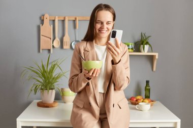 Çekici, gülümseyen, kahverengi saçlı, resmi takım elbiseli, mutfakta poz veren, kahvaltı yaparken görüntülü konuşan, blogu için yemek yerken selfi yapan bir kadın..