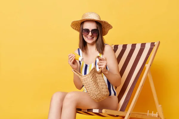 笑顔喜び女性身に着けているストライプ水着に座ってデッキチェア隔離上の黄色の背景に彼女の日焼け止めに袋 — ストック写真