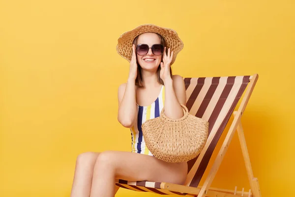 縞模様の水着を着たリラックスした女性の笑顔サングラスと帽子デッキチェアに座って黄色の背景に隔離された休暇を楽しんでビーチでリラックス — ストック写真