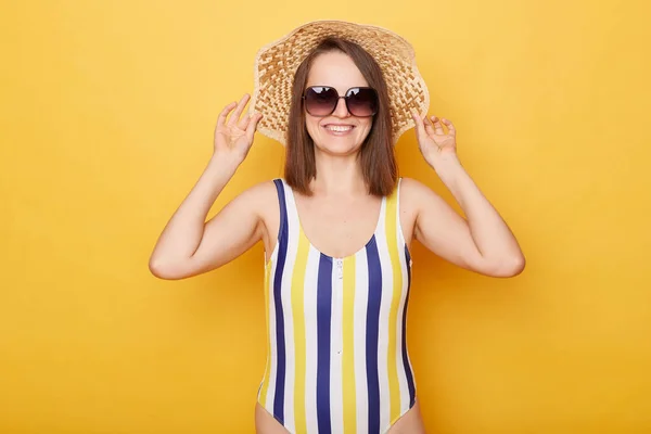 可愛いです笑顔喜びますCaucasian女性身に着けていますストライプ水着を身に着けています立って隔離された黄色の背景上に彼女の帽子楽しみますリゾートで休息 — ストック写真