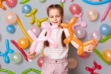 Gülümseyen mutlu bayan çocuk doğum günü partisinde oynuyor, örgülü şirin küçük kız gri duvara karşı duruyor renkli balonlarla süslenmiş..