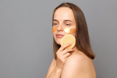 Güzellik portresi. Çekici, sakin, kahverengi saçlı, gri arka planda yalnız poz veren genç bir manken. Gözlerinin altında sünger tutan yamalar var. Dalgın yüzüyle başka yere bakıyor..