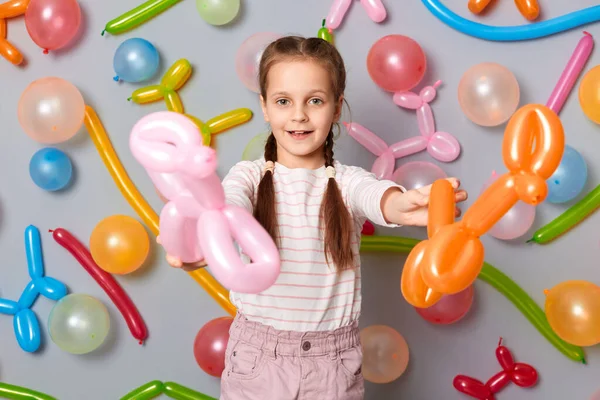 Take it. Having fun on party. Smiling cute little girl with braids standing against gray decorated wall, giving her multicolored balloons, looking at camera.