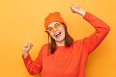 Heyecanlı, turuncu kazak giyen ve gözlüklü, sarı arka planda izole edilmiş kollarını kaldırmış, neşeli bir şekilde dans eden neşeli bir kadın..