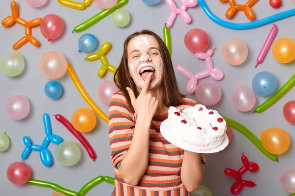 happy smiling woman with cake cream on face wearing striped dress standing against gray wall decorated with colorful balloons licking finger with birthday dessert.