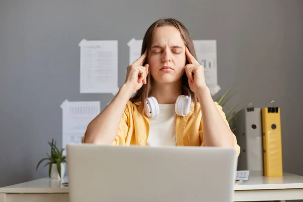 Young caucasian woman stressed and tired from work, holding fingers on temples, massaging, keeps eyes closed, suffering headache, working on laptop in office.