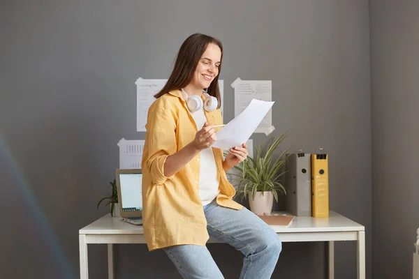 Young smiling employee woman in yellow jacket and jeans sitting on table in office holding paper documents working with charts and graphs.