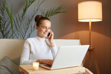 Beyaz kazaklı, kahverengi saçlı bir kadın dizüstü bilgisayara bakıyor ve uzaktan kumandalı akıllı telefondan konuşuyor. Evde kanepede oturuyor..