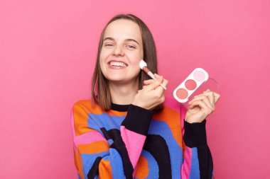 Makyaj yapan memnun bir kadın pembe arka planda izole edilmiş kozmetik fırça kullanıyor, kameraya gülümsüyor, pudra sürüyor..