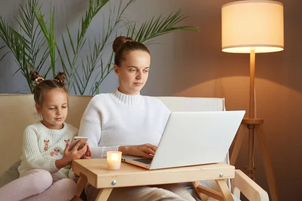 认真集中妈妈看笔记本电脑 坐在沙发上和妈妈坐在一起 用智能手机等妈妈完成她的工作任务 — 图库照片