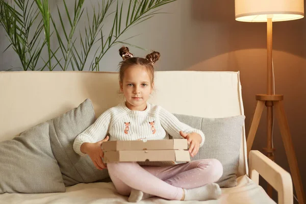 Tasty fast food snack. Cute little girl sitting on couch holding Pizza box at home, yummy meal, kid wearing casual clothing with hair buns enjoying delicious junk food looking at camera.