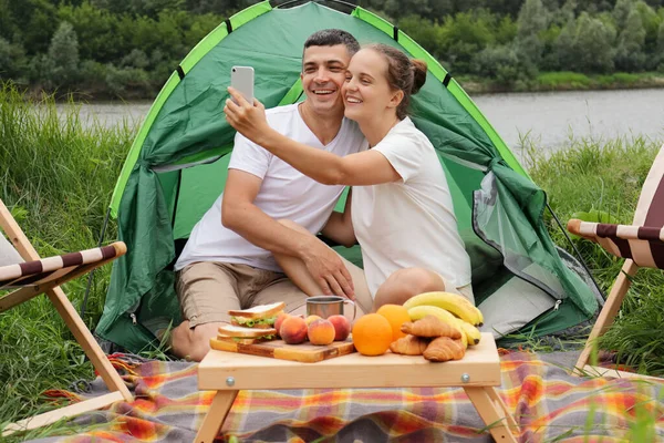 快乐的女人和男人穿着休闲装坐在河边的帐篷里 一边打电话一边享受大自然的休息 — 图库照片