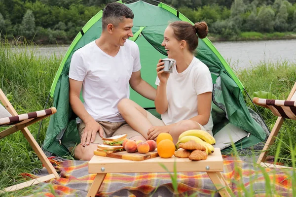 户外休闲 在自然界中休息 在河边露营 快乐的笑着的女人和穿着休闲装的男人 坐在地上搭帐篷 与人交流 享受假期 — 图库照片