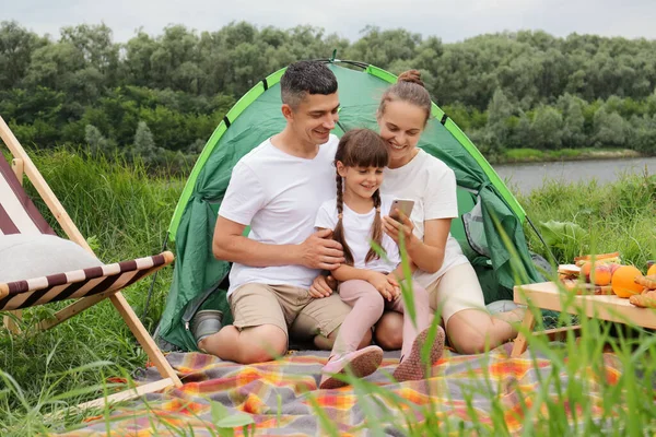 一家人坐在河边的地面上 在户外消磨时间 休闲活动 野外露营 在户外休息时使用智能手机的人 — 图库照片