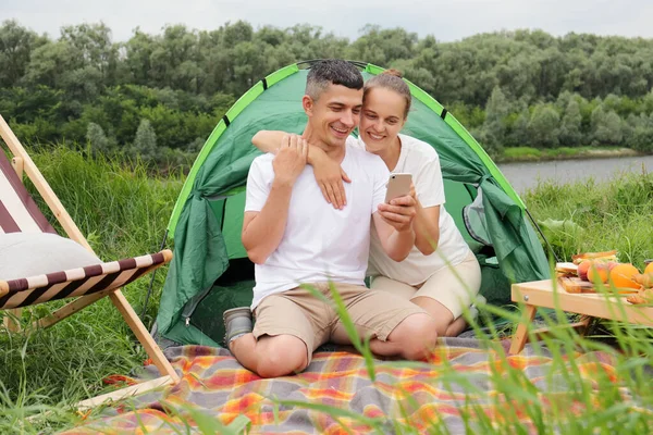 携帯電話を使って川の近くのテントに腰を下ろし 自然の中で休みを取りながらネットワークをチェックしている美しい女性とハンサムな男 — ストック写真