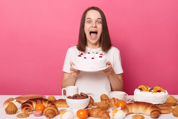 Overjoyed screaming brown haired woman in white t shirt sitting at table showing tasty cake breaking diet or having cheat meal isolated over pink background.