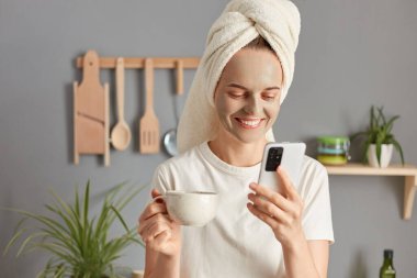 Neşeli gülümseyen genç bir kadın kafasında havluyla mutfakta poz veriyor cep telefonu kullanıyor kozmetoloji prosedürleri yapıyor sosyal ağları kontrol ediyor ve sabahları kahve ya da çay içiyor..