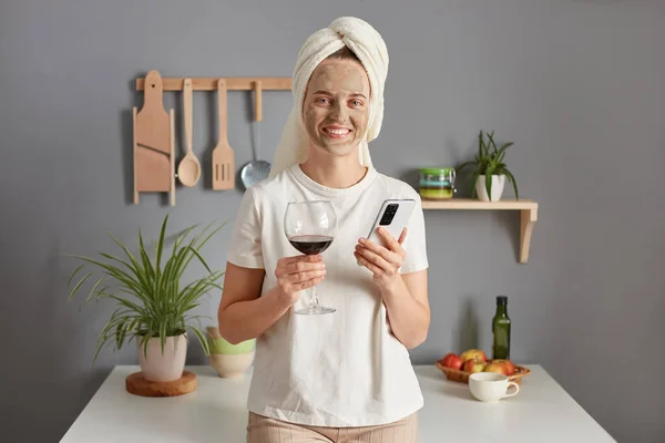 快乐而快乐的年轻女子头戴毛巾坐在厨房里 看着镜头 迷人地微笑着 用手机 — 图库照片