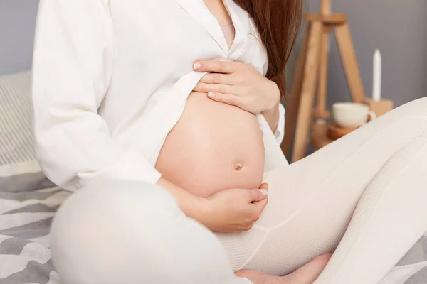 認識できない妊娠中の女性と一緒に座って彼女のおなかを保持し 白い服を着て休んで ベッドの上で屋内でリラックス赤ちゃんを期待 — ストック写真