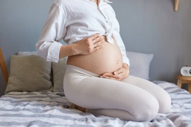 Sağlıklı hamilelik. Çocuk doğurmayı bekliyorum. Tanımlanamayan, beyaz elbiseli hamile bir kadın. Yatak odasında çıplak karnını tutarak oturuyor..