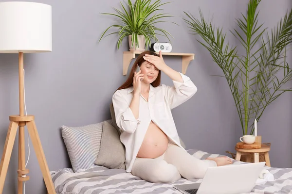 妊娠中の健康 電話相談 無病息災妊娠中の茶色の髪の白人女性がベッドに座って 頭痛と高温を持つ医師とスマートフォンで話す — ストック写真