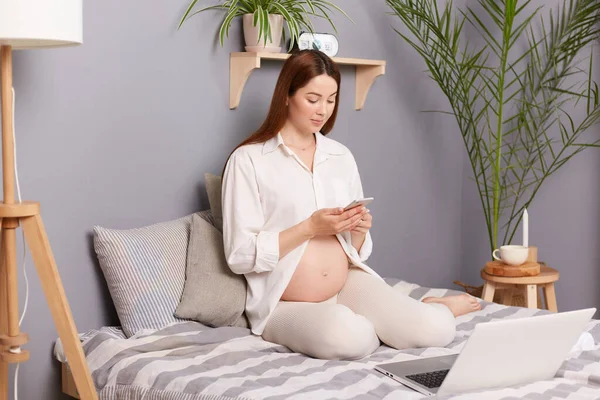 一个非常积极的怀孕棕色头发的高加索女人 她坐在床上 用手机打字和笔记本电脑上网 — 图库照片