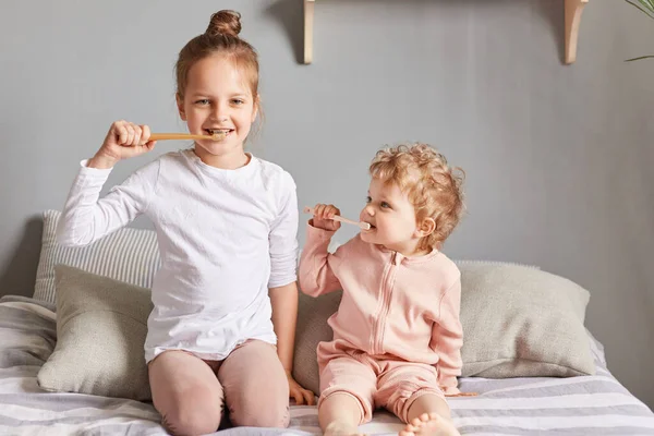 Daily routine hygiene habit. Baby girl and elder sister sitting on bed playing with toothbrush, smiling, siblings brushing teeth posing in bedroom.