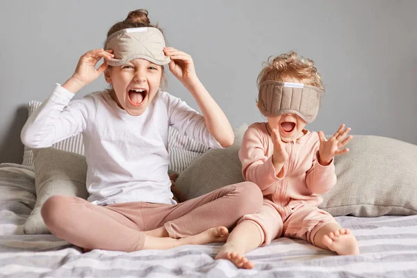 在卧室里玩耍的疯狂滑稽小女孩的画像 戴着眼罩的妹妹们在欢乐的气氛中醒来坐在床上尖叫着 — 图库照片