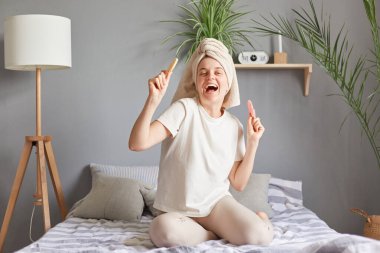 Neşeli, aşırı mutlu, beyaz havluyla mutlu bir kadın güzellik prosedürleri için yeni süngerler tutuyor ve hafta sonu duştan sonra yatakta dans ediyor..