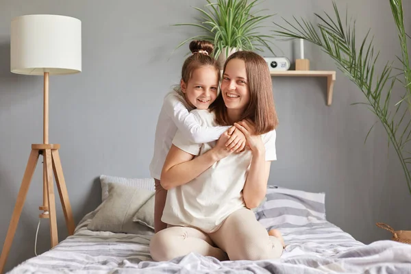 Stock image Joyful family in bedroom. Smiling brown haired mother and daughter sitting in bed at home happy kid hugging mommy enjoying weekend.