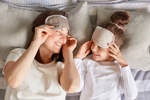 トップビューポートレートの茶色の髪の女性とともに彼女の娘で眠っているマスク嘘でベッドの中でお互いを見て笑顔と愛目覚めで週末の朝 — ストック写真