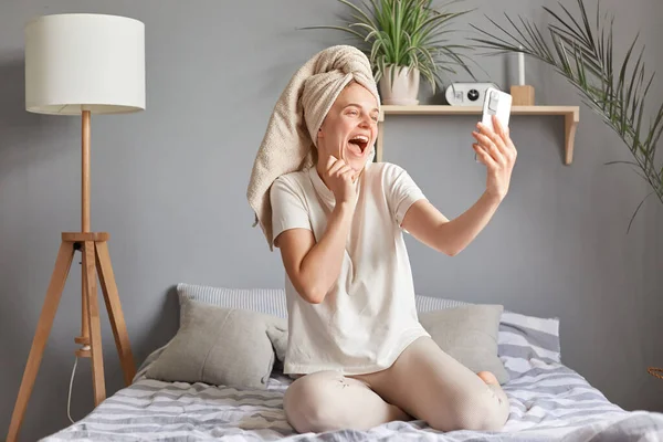 彼女のベッドに座って 自撮りを取り 手にスマートフォンを持ち 携帯電話でビデオ通話をしているタオルに包まれた魅力的な幸せな喜びの明るい女性 — ストック写真