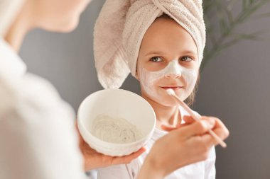 Tanımlanamayan anne ve küçük kızı evde birlikte poz veriyorlar. Yüzünde kremayla gülümseyen mutlu çocuk anne ailesinin güzellik tedavisine gülümsüyor..